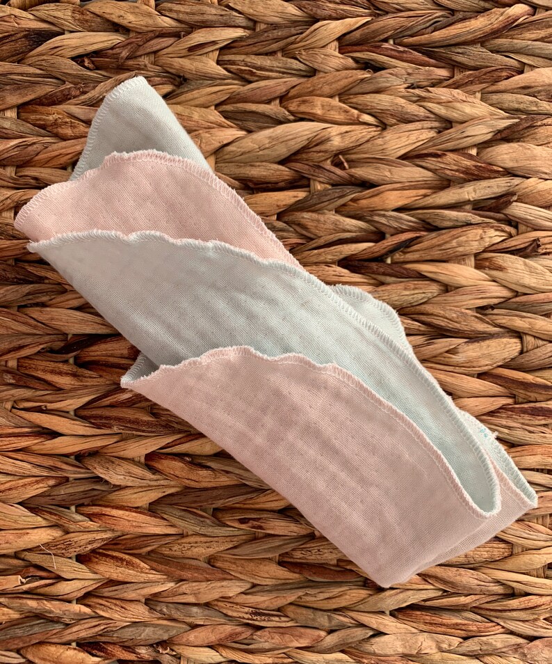 May include: Two folded cotton cloths, one light pink and one light blue, with scalloped edges. The cloths are folded and placed on a woven brown surface.