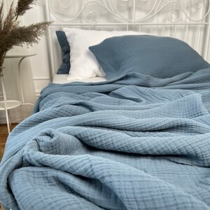 Pu&ograve; includere: Una morbida coperta in cotone testurizzato di colore blu &egrave; piegata su un letto bianco con un cuscino bianco e blu. La coperta &egrave; realizzata in un tessuto leggero e traspirante.
