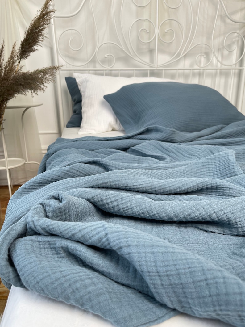 Pu&ograve; includere: Una morbida coperta in cotone testurizzato di colore blu &egrave; piegata su un letto bianco con un cuscino bianco e blu. La coperta &egrave; realizzata in un tessuto leggero e traspirante.