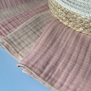 May include: A stack of four pink and beige muslin cloths with a white stitched edge. The cloths are folded and arranged on a light blue surface.