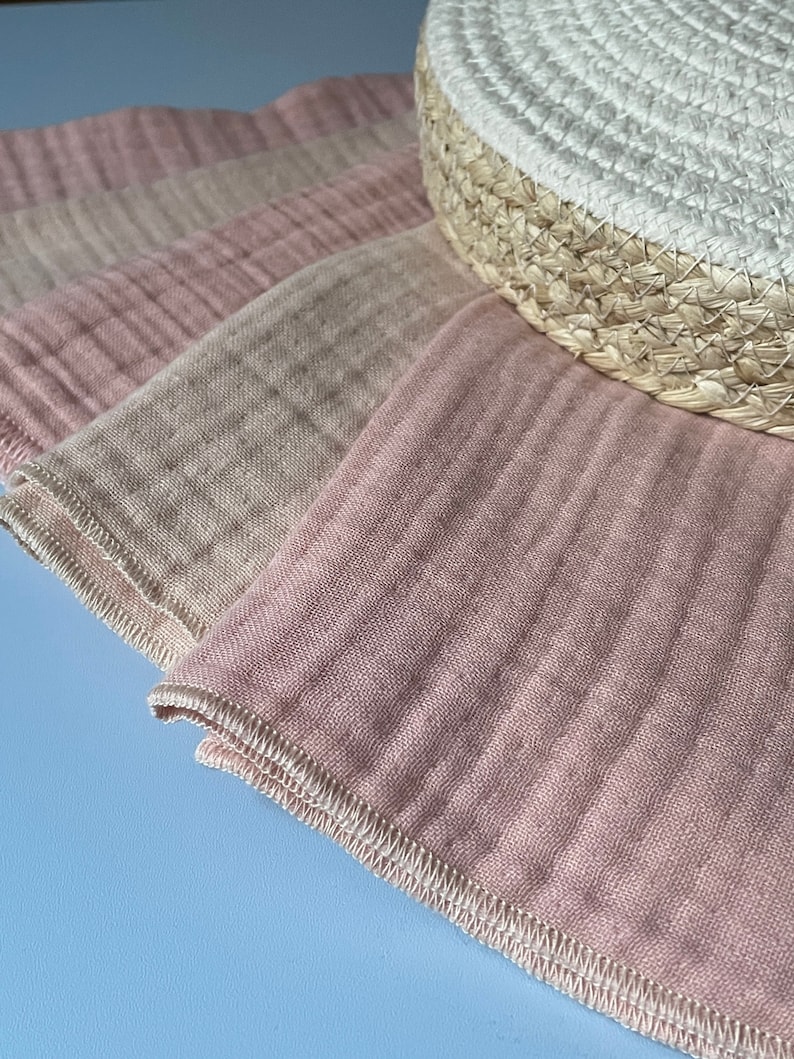 May include: A stack of four pink and beige muslin cloths with a white stitched edge. The cloths are folded and arranged on a light blue surface.