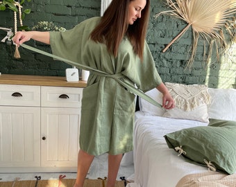 May include: A sage green linen robe with a self-tie belt. The robe is worn by a person standing in a bedroom with a white bed and a green brick wall.
