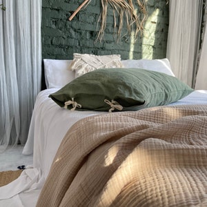 May include: A bed with a white linen duvet cover and a green linen pillow. The bed is made with a brown waffle weave throw blanket. The bed is in front of a green brick wall with a dried palm leaf hanging on the wall.