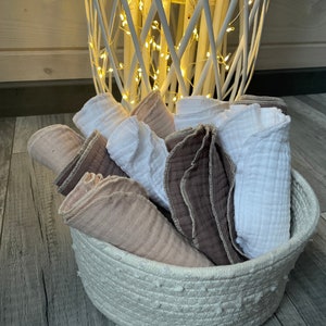 May include: A white woven basket filled with soft, white and pink muslin cloths. The cloths are folded and stacked in the basket.