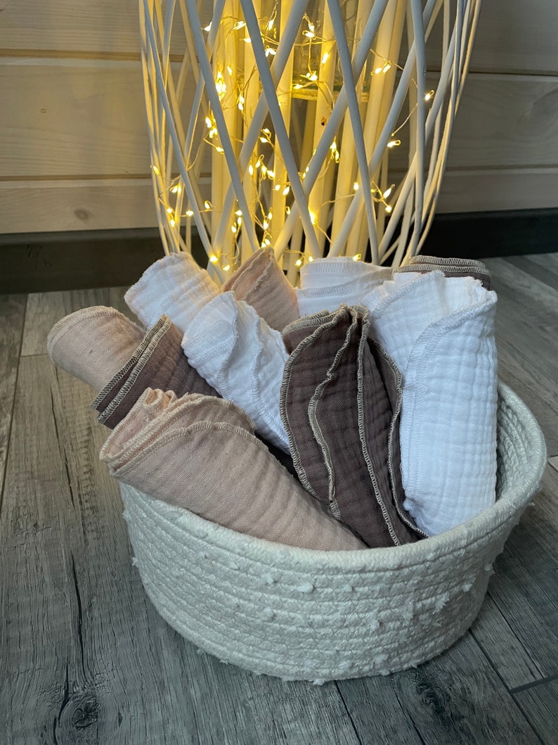 May include: A white woven basket filled with soft, white and pink muslin cloths. The cloths are folded and stacked in the basket.