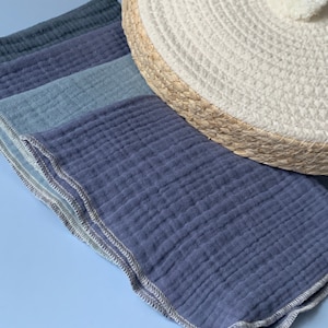 May include: A stack of four blue and gray muslin cloths with a white stitched border. The cloths are folded and arranged on a light blue surface.