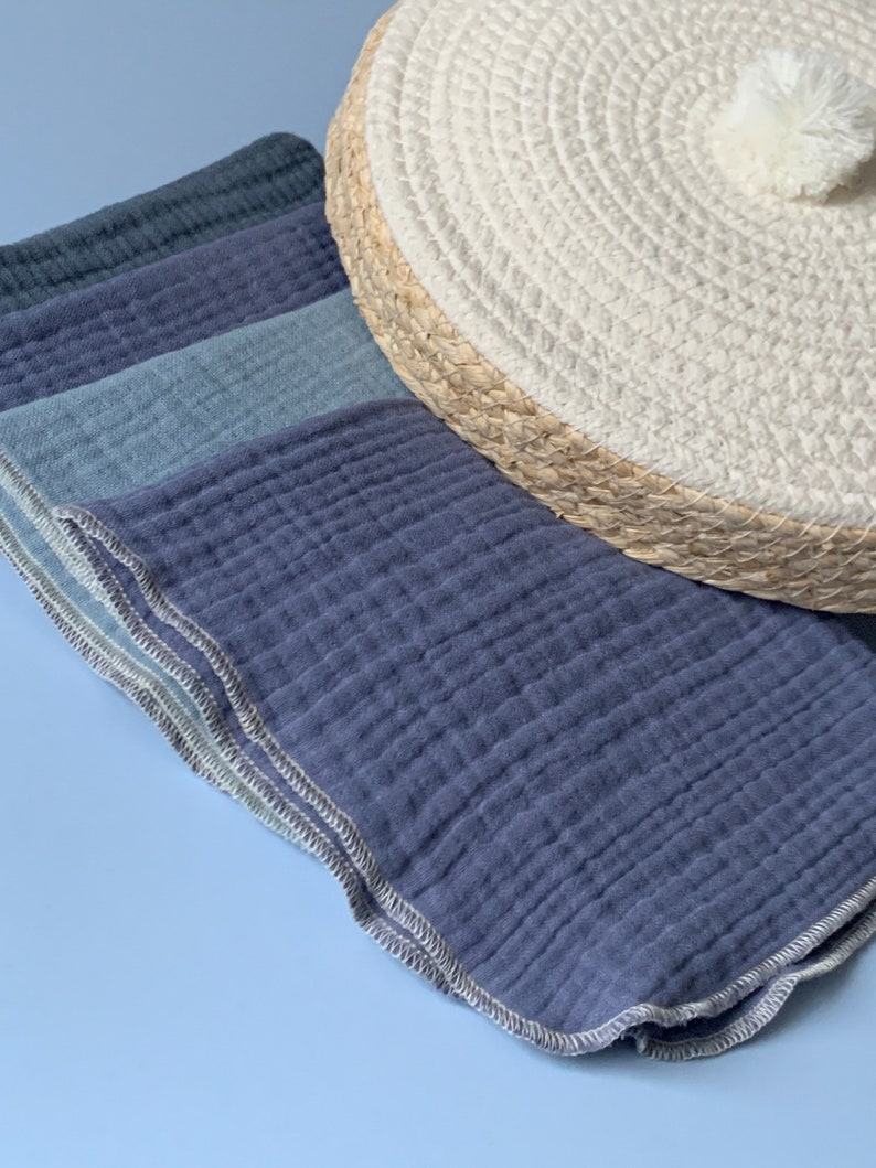 May include: A stack of four blue and gray muslin cloths with a white stitched border. The cloths are folded and arranged on a light blue surface.