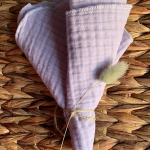 May include: A set of three lavender-colored muslin cloths folded and tied together with twine. The cloths have a white stitched edge and are displayed on a woven wicker surface.