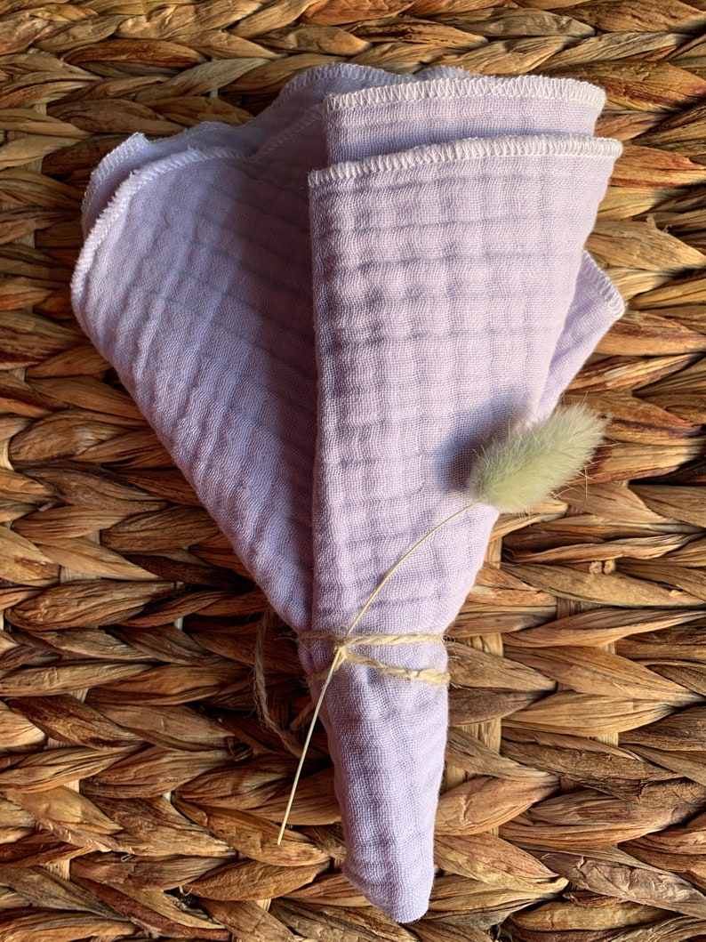May include: A set of three lavender-colored muslin cloths folded and tied together with twine. The cloths have a white stitched edge and are displayed on a woven wicker surface.