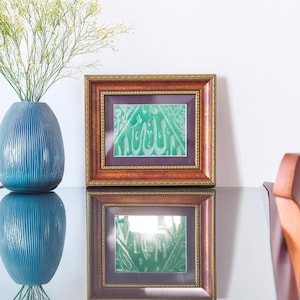 May include: A blue vase with vertical lines holds white flowers, next to a framed artwork with green calligraphy. The artwork is in a brown frame with a gold trim. The artwork is reflected on a glass surface.