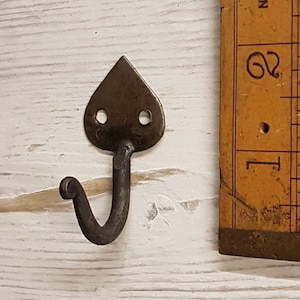 Pode incluir: Um gancho de metal preto com uma parte superior em forma de coração. O gancho mede aproximadamente 5 cm de comprimento e tem dois pequenos orifícios na parte superior. O gancho é mostrado ao lado de uma régua.