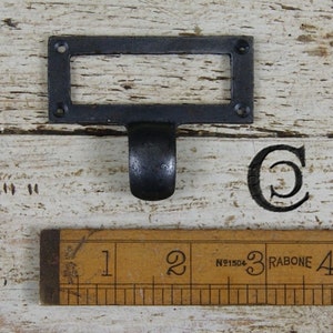 May include: A black metal label holder with a pull handle, suitable for drawers or cabinets. The label holder has a rectangular frame with four screw holes. A wooden ruler with centimetre markings is below the label holder.