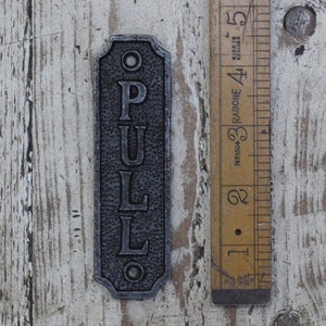 Può includere: Un cartello in metallo nero con la parola "PULL" scritta verticalmente in lettere bianche. Il cartello è alto circa 12,7 cm e ha una parte superiore e inferiore leggermente arrotondata.