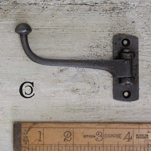 Peut inclure: Un crochet en métal noir avec un sommet arrondi et une base à charnière. Le crochet est monté sur un mur blanc. Le crochet mesure environ 10 cm de long.