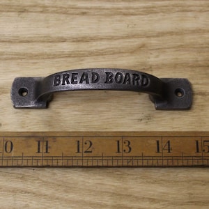 Op de afbeelding: Een zwart gietijzeren broodplank handvat met de woorden "Bread Board" gegraveerd op de voorkant. Het handvat is ongeveer 13 inch lang.