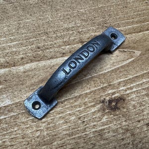 Peut inclure: Une poignée en métal gris foncé avec le mot "LONDON" en relief au centre incurvé. La poignée a une base rectangulaire avec un trou à chaque extrémité pour le montage. La poignée est posée sur un fond en bois.