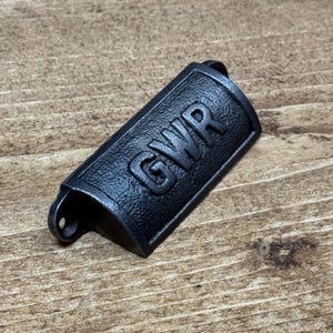 May include: A black metal drawer pull with the letters "GWR" embossed on the front. The pull is rectangular with rounded edges and a small hole on one end. It rests on a wooden surface.