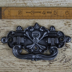 May include: A dark gray metal drawer pull with a skull and crossbones design. The handle has an ornate, decorative shape with a curved bottom and a rectangular section for the drawer hardware. A wooden ruler is in the background.