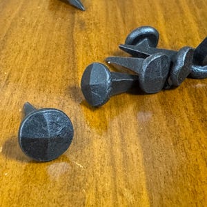 Puede incluir: Primer plano de varias cabezas de clavos decorativas negras con un diseño redondo facetado. Los clavos están dispersos sobre una superficie de madera. Los clavos tienen un aspecto rústico y vintage, adecuados para proyectos de carpintería o artesanía.