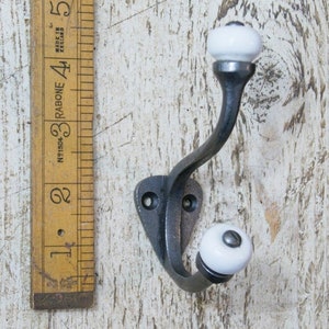 May include: A black metal hook with two white ceramic knobs. The hook is mounted on a wooden surface.