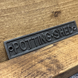 May include: A rectangular, dark grey metal sign with the words "POTTING SHED" in raised lettering. The sign has two holes for mounting and is set against a wooden background.