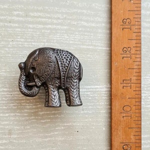 Può includere: Piccola statuetta di elefante in metallo in stile antico con motivi incisi dettagliati. L'elefante è di colore argento tenue e misura circa 4 cm di lunghezza. Un righello di legno è accanto all'elefante.