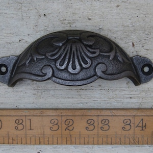 Op de afbeelding: Een zwarte gietijzeren ladegreep met een decoratief bloemmotief. De greep is ongeveer 7,5 cm breed.