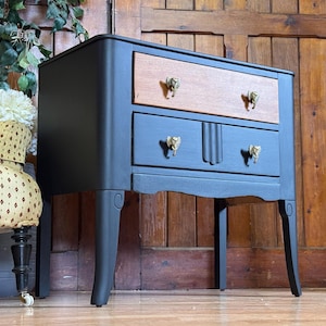 May include: A two-drawer nightstand painted in black and blue with gold-coloured elephant head drawer pulls. The top drawer is natural wood. The nightstand has curved legs and a decorative apron.