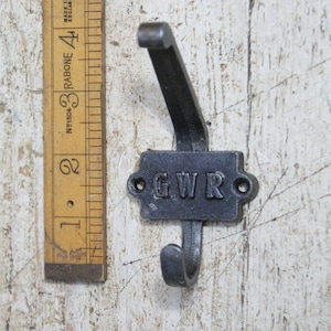 Pode incluir: Um gancho de ferro fundido preto com uma placa retangular que diz "GWR" em letras em relevo. O gancho está montado em uma superfície de madeira branca.