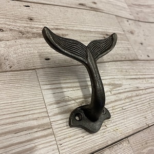 Może przedstawiać: Czarny, żeliwny hak ścienny w kształcie ogona wieloryba. Hak ma rustykalne wykończenie.