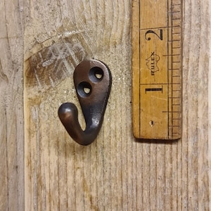 Peut inclure: Un petit crochet en métal de couleur bronze avec deux trous de vis et un crochet incurvé. Le crochet mesure environ 5 cm de long et est monté sur une surface en bois vieilli. Une règle vintage est à côté du crochet.