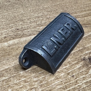 May include: A dark gray metal bottle opener with the word "LINER" embossed on the front. The opener has a textured surface and a small hole on one end. The opener is on a wooden surface.