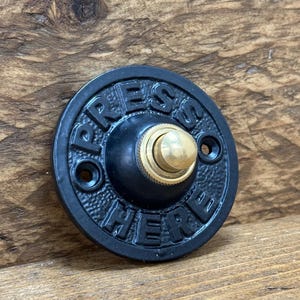 Può includere: Un pulsante del campanello nero e circolare con le parole "PRESS HERE" in rilievo sul bordo. Il pulsante ha un centro color ottone e due piccoli fori per il montaggio. Il campanello è in metallo.