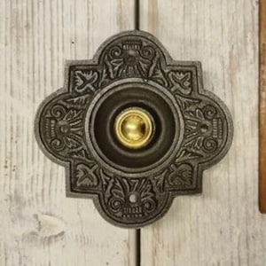 May include: Decorative cast iron doorbell button with a dark grey finish and a gold-coloured button. The button has a four-lobed design with floral and scrollwork details. It is mounted on a light-coloured wooden surface.