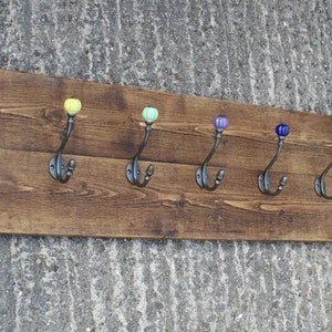 Pode incluir: Um cabide de madeira com oito ganchos, cada um com um botão de cerâmica de cor diferente. Os botões são vermelho, amarelo, verde, verde claro, roxo, azul, azul claro e laranja.