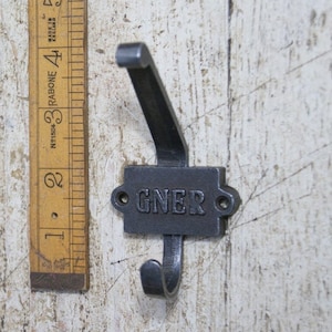 Pode incluir: Gancho preto de ferro fundido com uma placa retangular que diz "GNER" em letras em relevo. O gancho está montado em uma parede de madeira branca.
