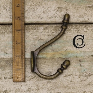 May include: Antique brass hook with a decorative acorn finial on each end. The hook is mounted on a wooden surface.