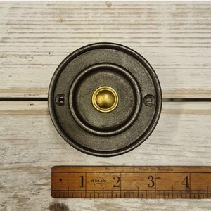 Könnte beinhalten: Ein runder, dunkelgrauer Metall-Türklingelknopf mit Messingmitte. Der Knopf hat ein kreisförmiges Design mit einem erhabenen Außenring und zwei Schraubenlöchern. Ein Holzlineal befindet sich im Bild.