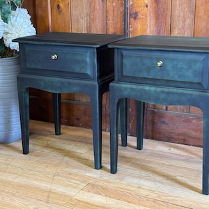 May include: A pair of dark teal bedside tables, each with a single drawer and a gold-coloured knob. The tables have a rectangular top and are supported by four curved legs. The background features a wooden door and a vase with white flowers.