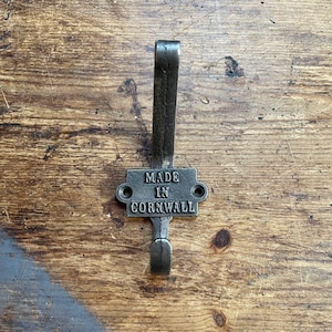 Pode incluir: Um gancho de metal preto com uma placa retangular que diz "MADE IN CORNWALL". O gancho está montado em uma superfície de madeira.