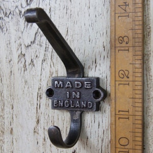 Pode incluir: Um gancho de parede de metal escuro com um gancho inferior curvo e uma placa retangular que diz "MADE IN ENGLAND". O gancho é montado em uma superfície de madeira branca desgastada. Uma régua de madeira está ao lado do gancho.