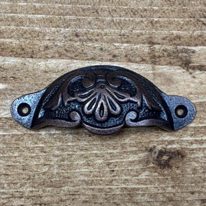 May include: An ornate drawer pull in a dark bronze finish. The metal handle has a symmetrical design with a central floral motif and two mounting holes. The hardware is set against a wooden background.