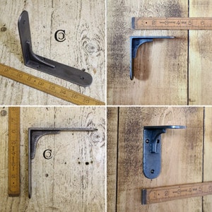 May include: Four black metal shelf brackets of different sizes and shapes. The brackets are shown against a wooden background with a ruler for scale.