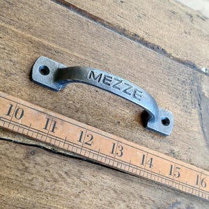 May include: A silver metal cabinet pull with the word "MEZZE" embossed on the front. The pull is about 4 inches long and is shown next to a ruler.