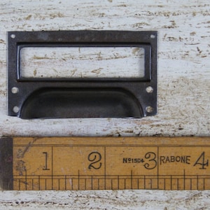 May include: A black metal drawer pull with a rectangular label holder. The pull is approximately 3 inches long.