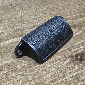 May include: A dark gray metal bottle opener with the text "COVENT GARDEN LONDON" embossed on the front. The opener has a textured surface and a small hole for hanging. It rests on a wooden surface.