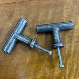 Könnte beinhalten: Zwei silberne T-förmige Schrankknöpfe mit Schrauben. Die Knöpfe haben eine abgenutzte Oberfläche.