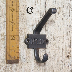 Pode incluir: Um gancho de casaco de ferro fundido preto com um único gancho e uma placa retangular com a palavra "NYMR" gravada em relevo.
