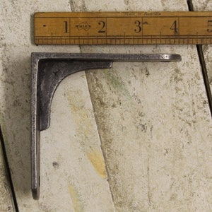 May include: A dark gray metal shelf bracket with a right-angle design. The bracket is positioned next to a wooden ruler with black markings. The ruler is marked in inches, with the bracket's length appearing to be approximately 4 inches.