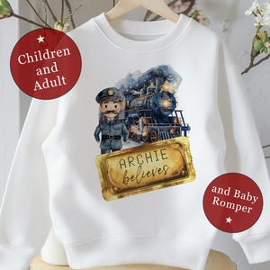 May include: White sweatshirt featuring a watercolor illustration of a train, a conductor, and the text "ARCHIE believes." The design includes blue, black, and gold accents. The sweatshirt is suitable for children and adults.